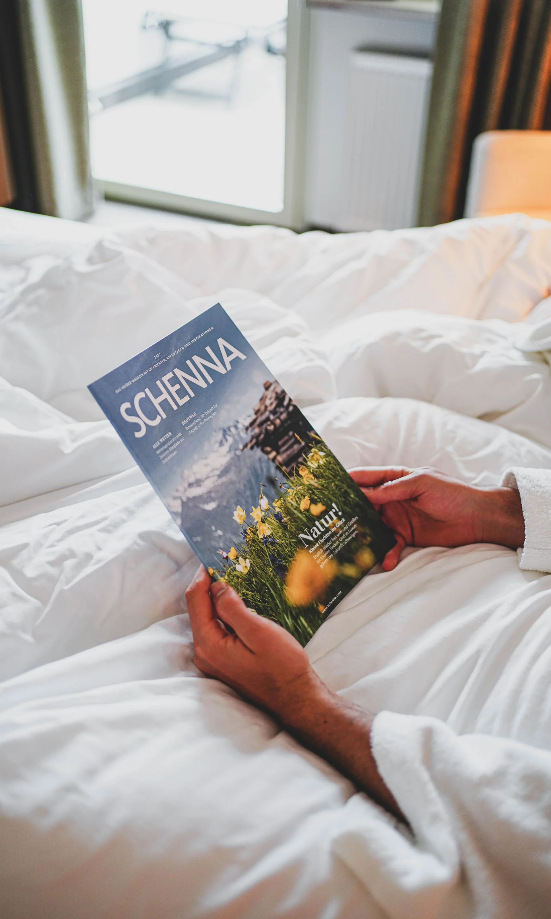 Euer Hotel in Schenna für Momente in #echtzeit Person hält Broschüre Schenna auf einem weißen Bett