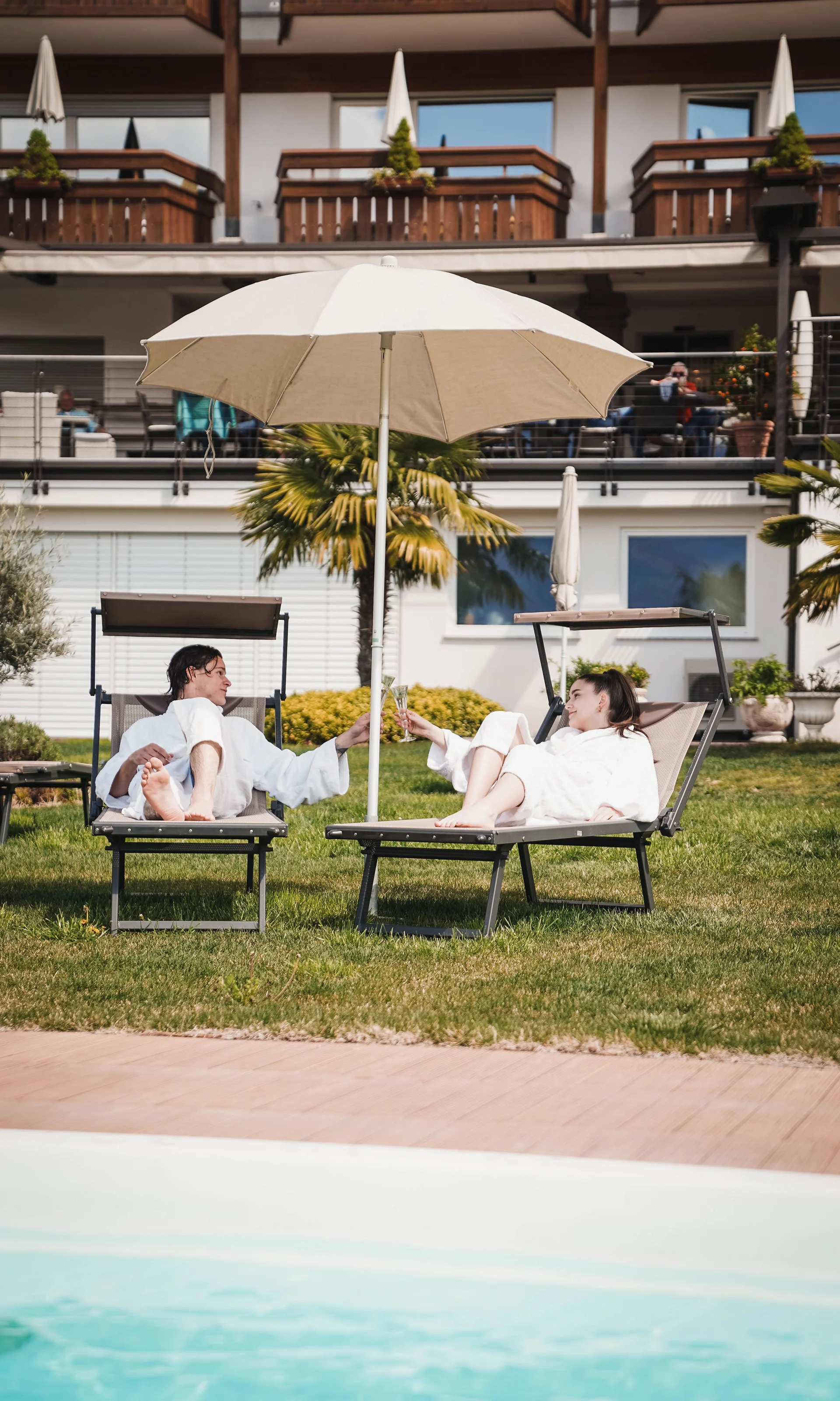 Paar in Bademänteln stößt bei Pool auf Liegestühlen an