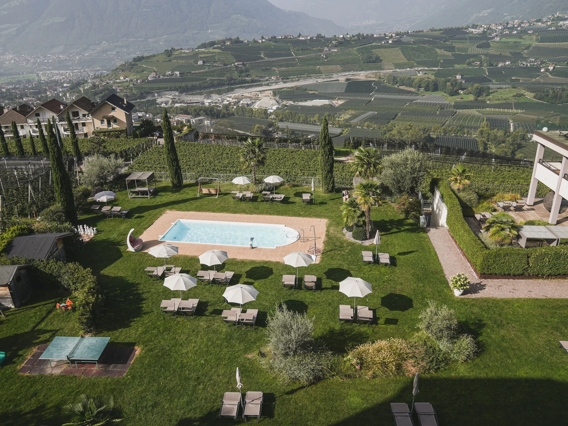 Hotelgarten mit Pool, Sonnenschirmen und Liegestühlen vor Weinbergen und Bergen