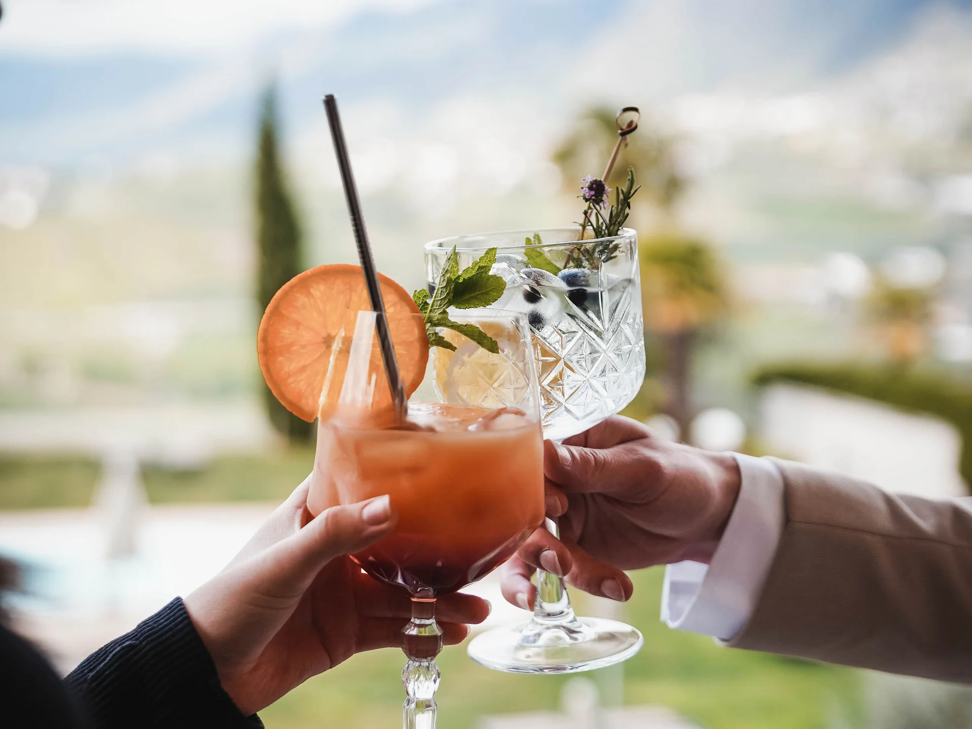 Euer Hotel in Schenna für Momente in #echtzeit Zwei Hände stoßen mit Cocktails mit Reifen und Kräutern am Balkon an