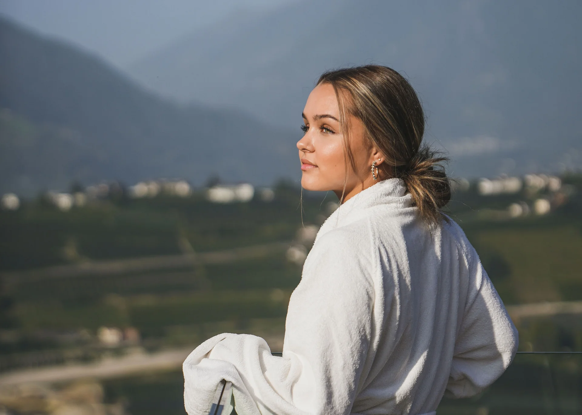 Frau im weißen Bademantel blickt auf Berglandschaft von Balkon aus