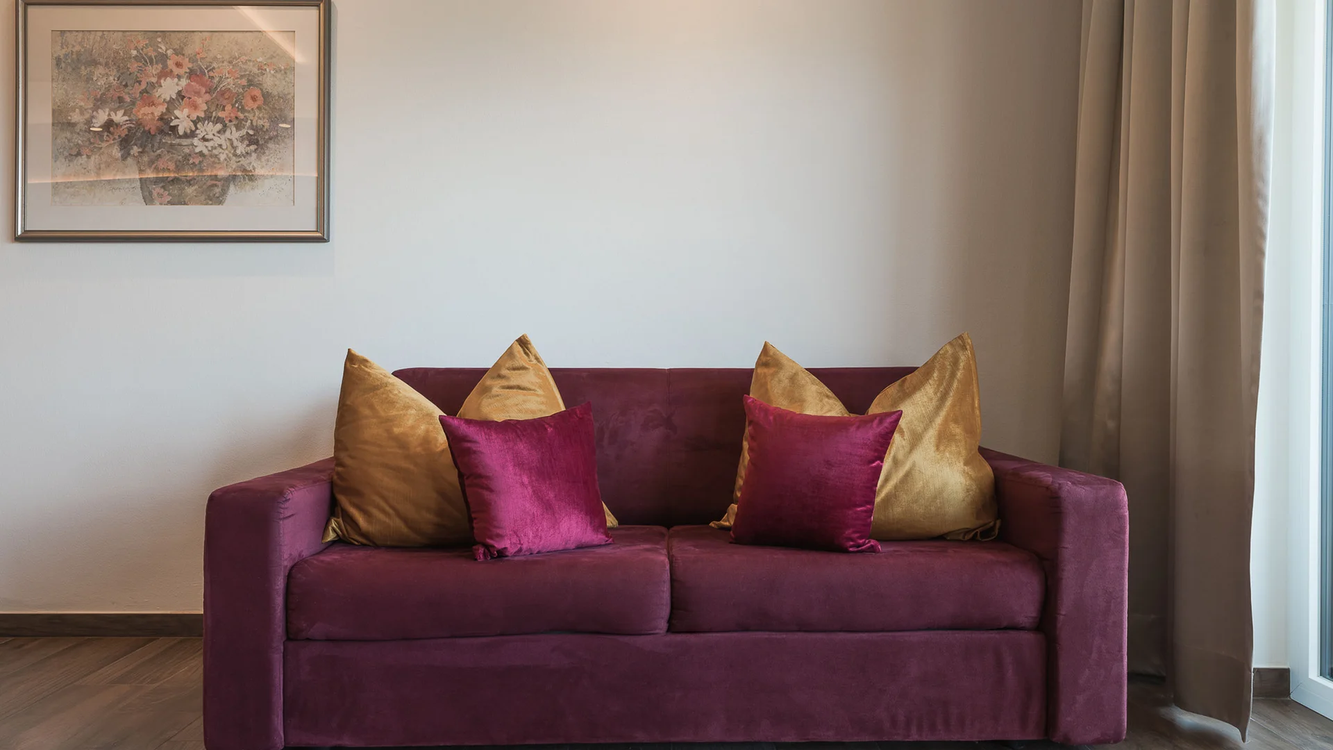 Impressionen in #echtzeit Lila Sofa mit goldenen und violetten Kissen vor weißer Wand mit Blumenbild